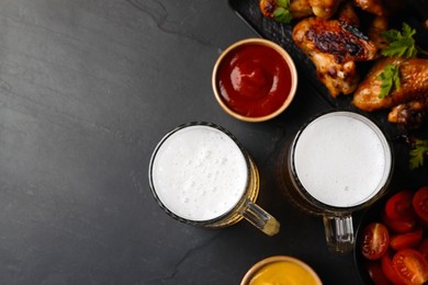 Photo of Mugs with beer, delicious baked chicken wings and sauces on dark grey table, flat lay. Space for text