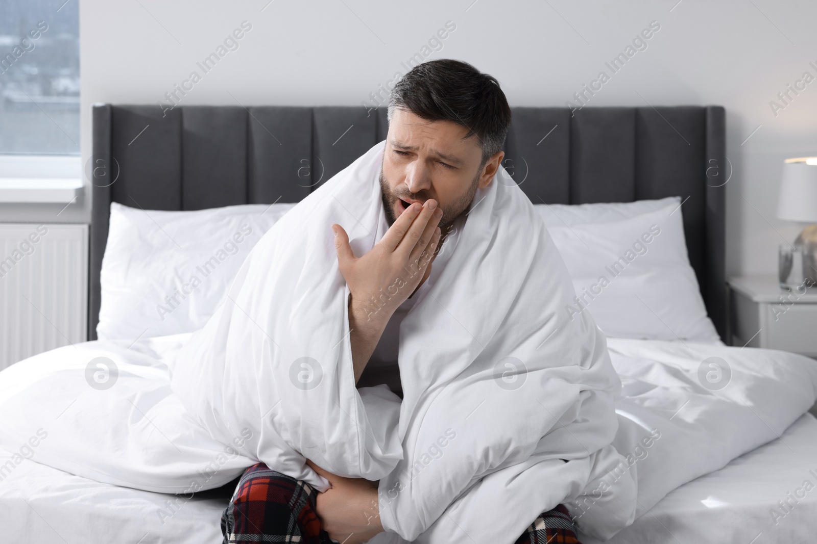 Photo of Sleepy man wrapped in blanket yawning on soft bed at home