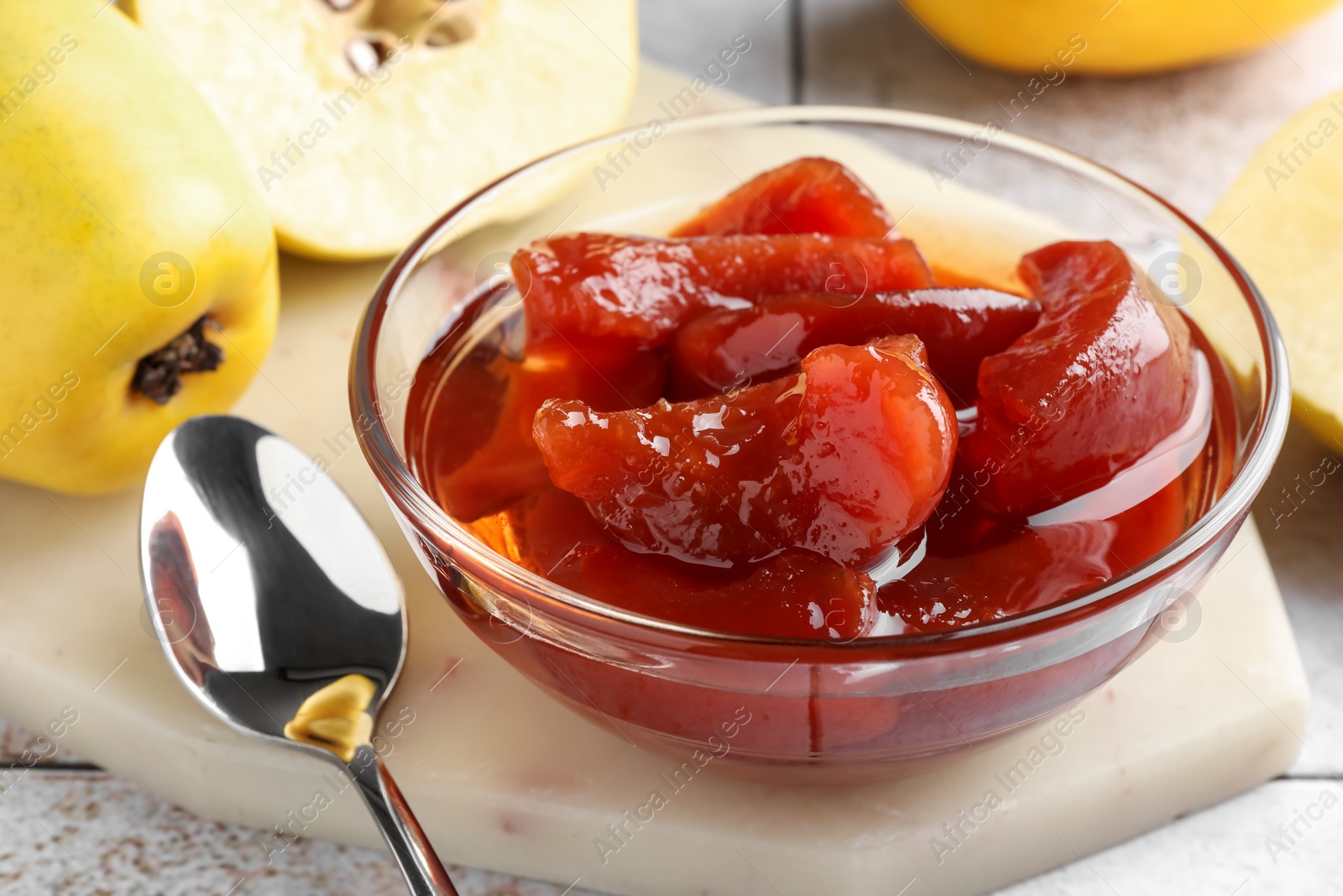 Photo of Tasty homemade quince jam in bowl, spoon and fruits on tiled table, closeup