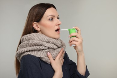 Woman with scarf using throat spray on grey background, space for text