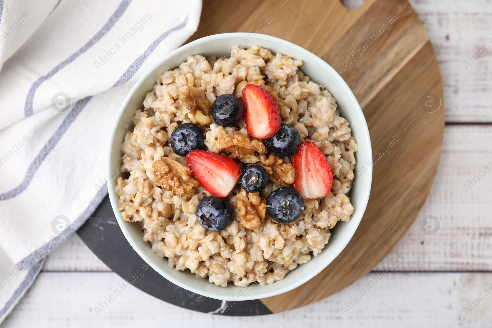 Photo of Tasty oatmeal with strawberries, blueberries and walnuts in bowl on white wooden table, top view