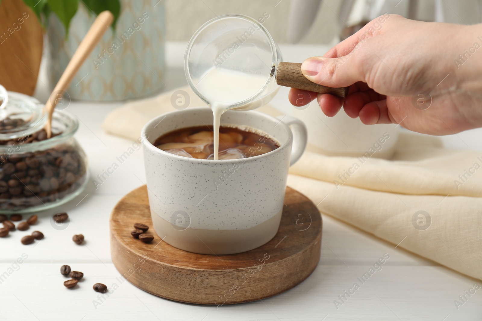 Photo of Woman pouring milk into cup with coffee at white wooden table, closeup