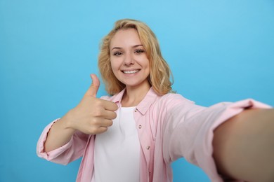 Photo of Happy young woman showing thumb up gesture while taking selfie on light blue background