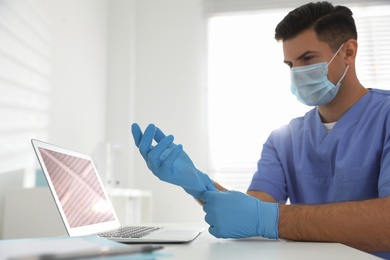 Doctor in protective mask putting on medical gloves at table in office