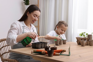 Mother with her daughter spraying water onto vegetable seeds in pots at wooden table indoors