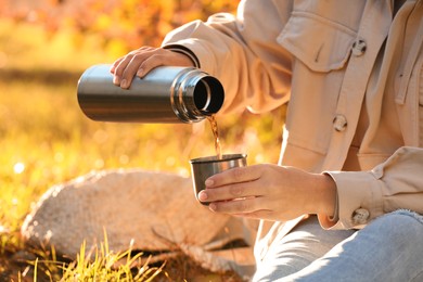 Photo of Picnic time. Woman pouring tea from thermos into cup lid on green grass outdoors, closeup