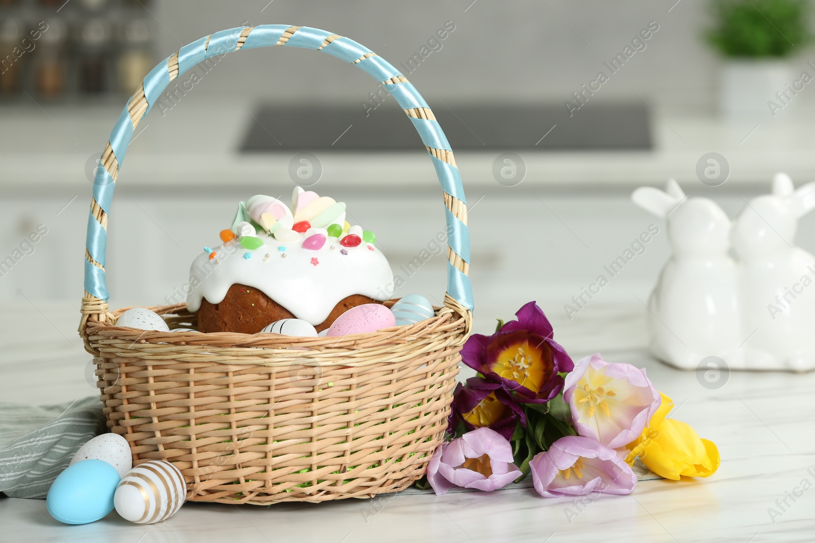 Photo of Easter basket with painted eggs and tasty cake near flowers on white marble table indoors
