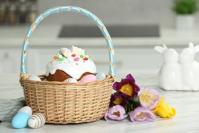 Easter basket with painted eggs and tasty cake near flowers on white marble table indoors