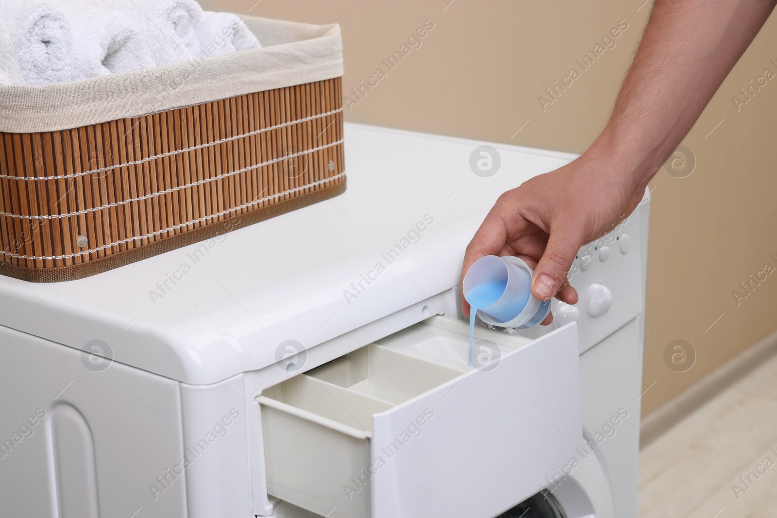 Photo of Man pouring fabric softener from cap into washing machine indoors, closeup