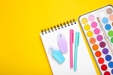 Watercolor palette, notebook and colorful markers on yellow background, flat lay. Space for text