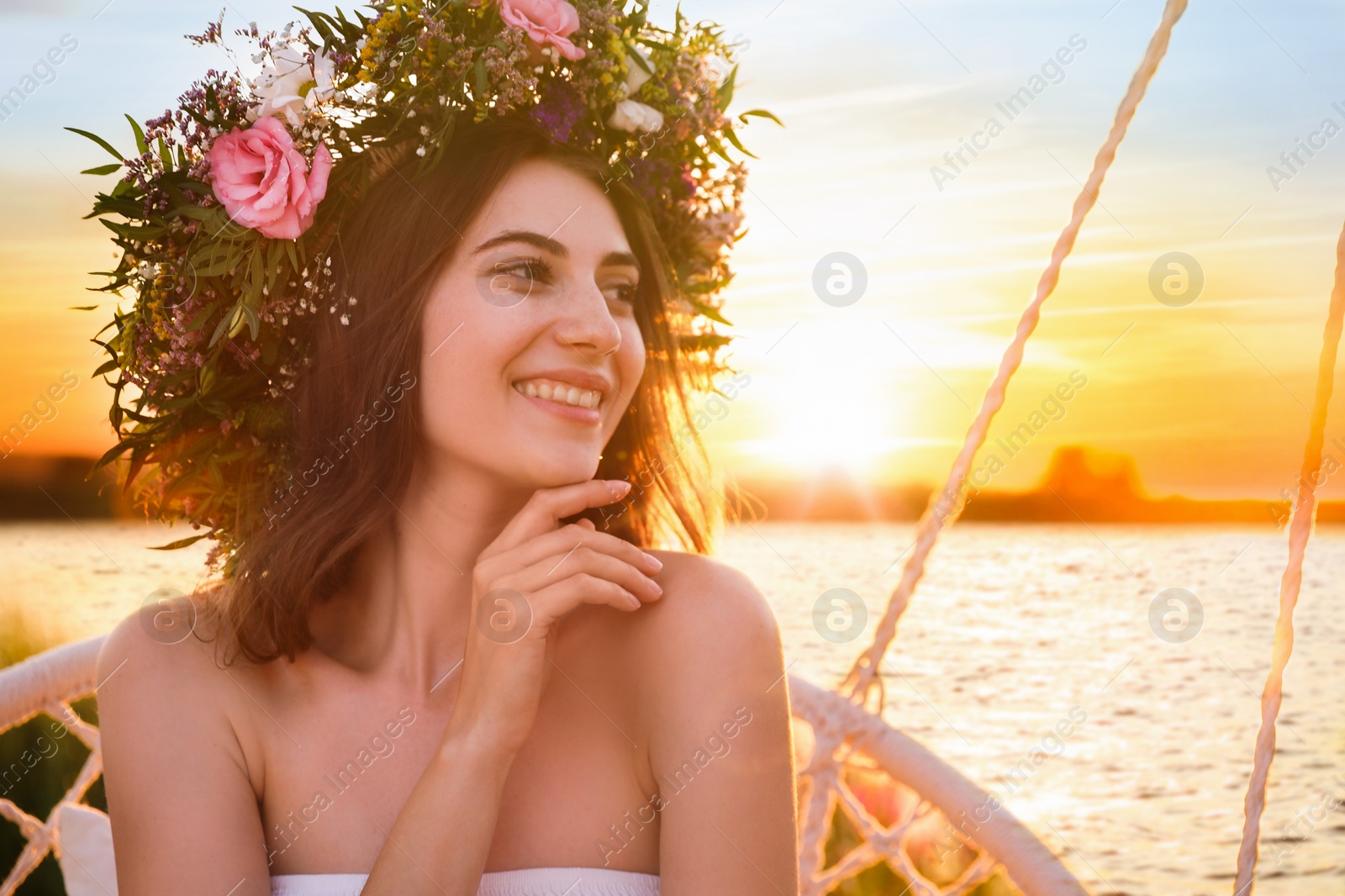 Photo of Young woman wearing wreath made of beautiful flowers on swing chair outdoors at sunset