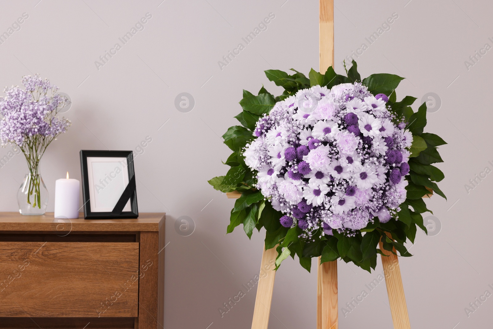 Image of Wreath of violet and white flowers, photo frame with black ribbon in room. Funeral attributes