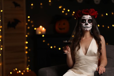 Photo of Young woman in scary bride costume with sugar skull makeup and glass of wine against blurred lights indoors, space for text. Halloween celebration