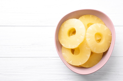 Delicious canned pineapple rings in bowl on white wooden table, top view. Space for text