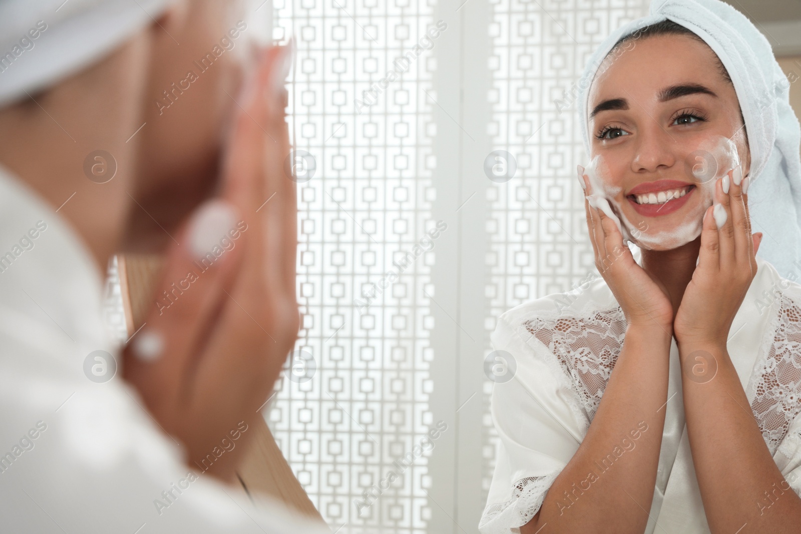 Photo of Beautiful young woman applying cleansing foam onto face near mirror in bathroom. Skin care cosmetic