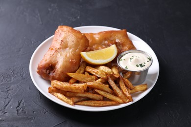 Photo of Tasty fish, chips, lemon and sauce on black table