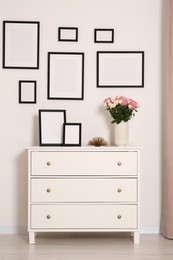 Empty frames hanging on white wall and chest of drawers with flowers in room