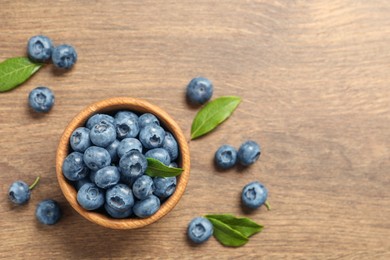 Photo of Bowl of tasty fresh blueberries with green leaves on wooden table, flat lay. Space for text