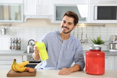 Photo of Young man holding bottle of protein shake at table with ingredients in kitchen