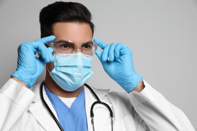 Doctor in protective mask, glasses and medical gloves against light grey background