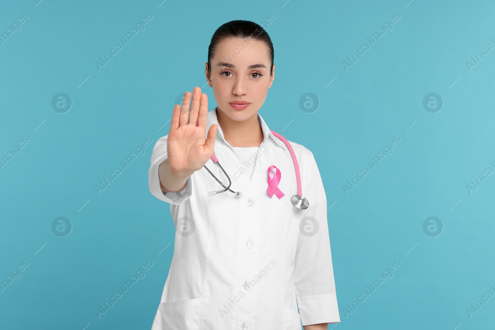 Photo of Mammologist with pink ribbon showing stop gesture on light blue background. Breast cancer awareness