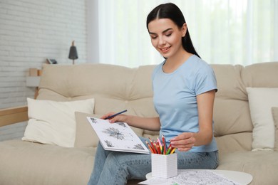Photo of Young woman with antistress coloring page taking pencil on sofa in living room
