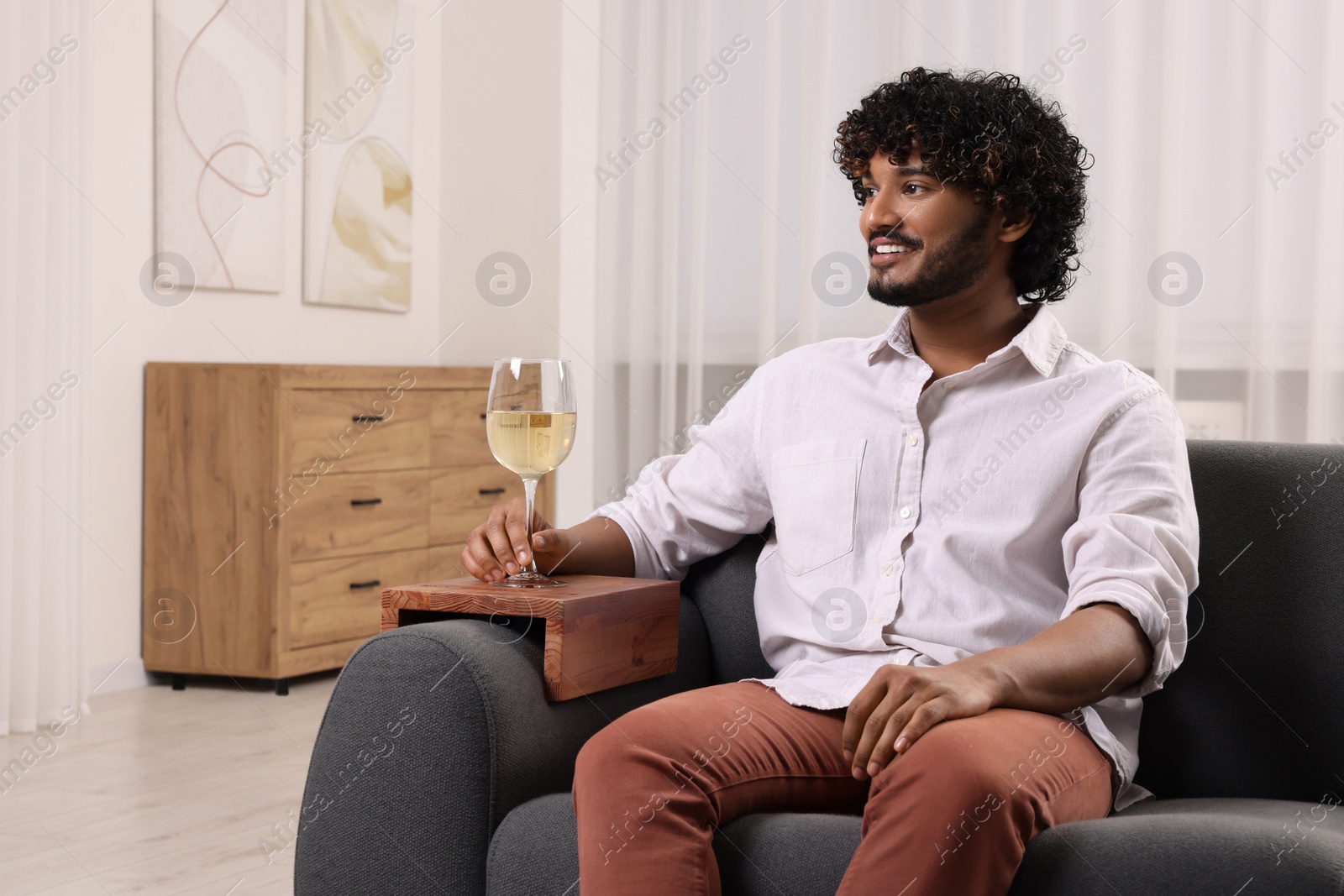 Photo of Happy man holding glass of wine on sofa armrest wooden table at home, space for text