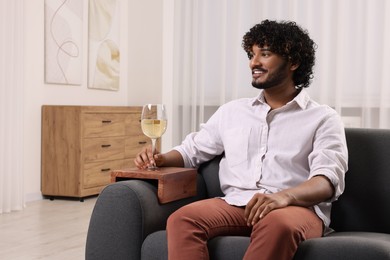 Happy man holding glass of wine on sofa armrest wooden table at home, space for text