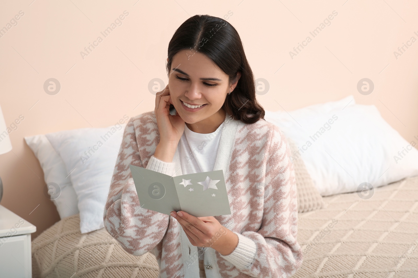 Photo of Young woman with greeting card on bed in room, space for text