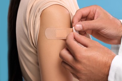 Doctor sticking plaster on woman's arm after vaccination against light blue background, closeup