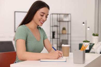 Photo of Young woman writing in notebook at white table indoors