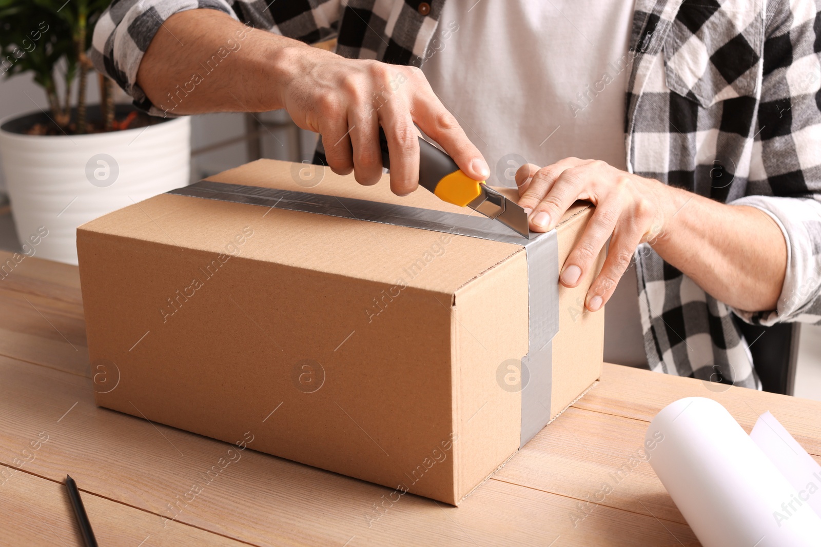 Photo of Man using utility knife to open parcel at wooden table indoors, closeup
