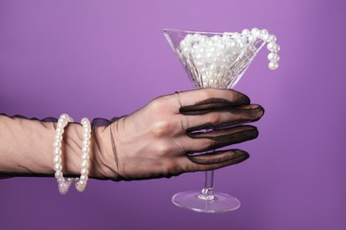 Woman in elegant gloves holding beautiful martini glass with pearls on purple background, closeup