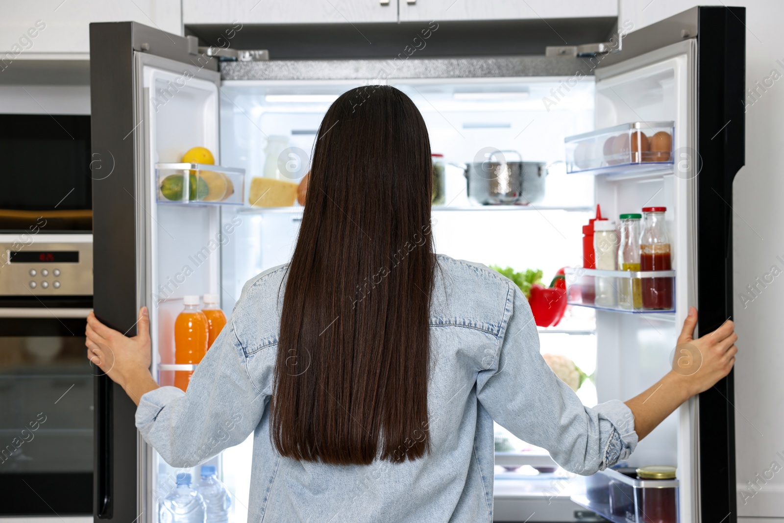 Photo of Young woman near modern refrigerator in kitchen, back view