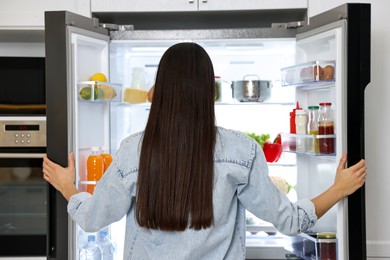 Young woman near modern refrigerator in kitchen, back view