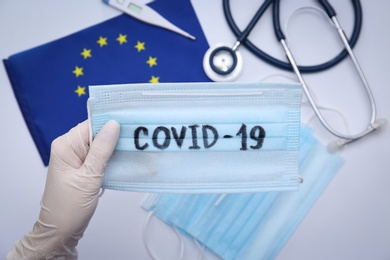 Photo of Doctor holding mask with word COVID-19 above medical items and European Union flag, closeup