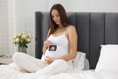 Photo of Pregnant woman with ultrasound picture of baby on bed indoors