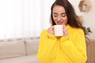 Photo of Beautiful young woman in stylish warm sweater holding cup of drink at home, space for text