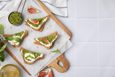 Tasty bruschettas with cream cheese, pesto sauce, figs and fresh basil on white tiled table, flat lay. Space for text