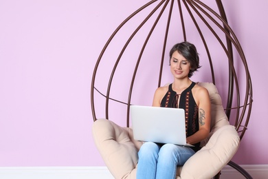Young woman with modern laptop sitting in hanging chair near color wall