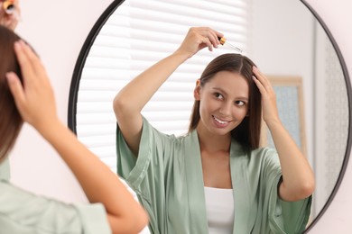 Photo of Beautiful woman applying serum onto hair near mirror indoors