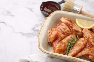 Photo of Fresh marinade, basting brush, raw chicken wings, rosemary and lemon on white marble table, closeup. Space for text