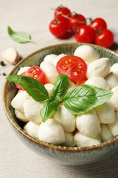 Photo of Delicious mozzarella balls, tomatoes and basil leaves in bowl on white wooden table, closeup