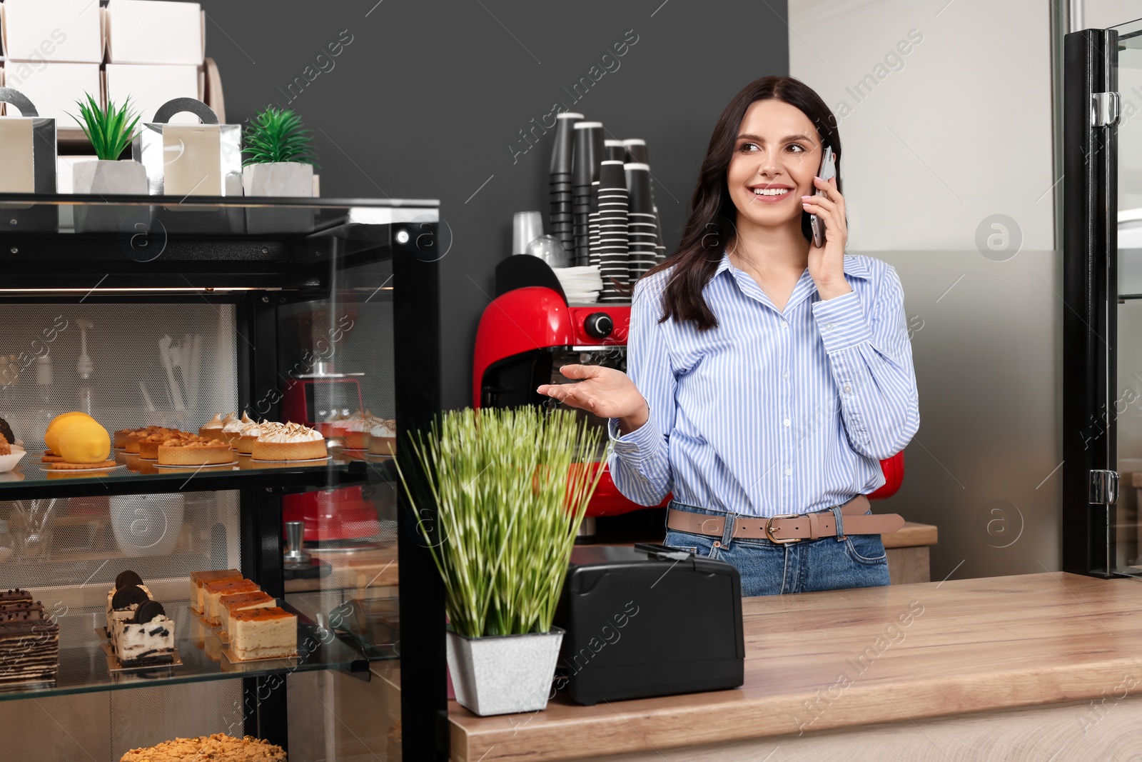 Photo of Business owner in her cafe. Woman talking on phone near showcase with pastries