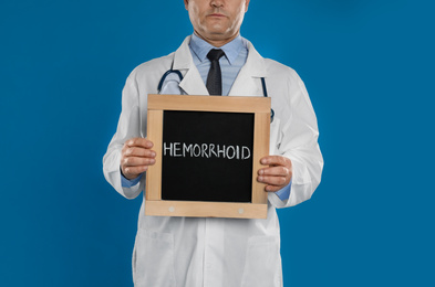 Doctor holding blackboard with word HEMORRHOID on blue background, closeup