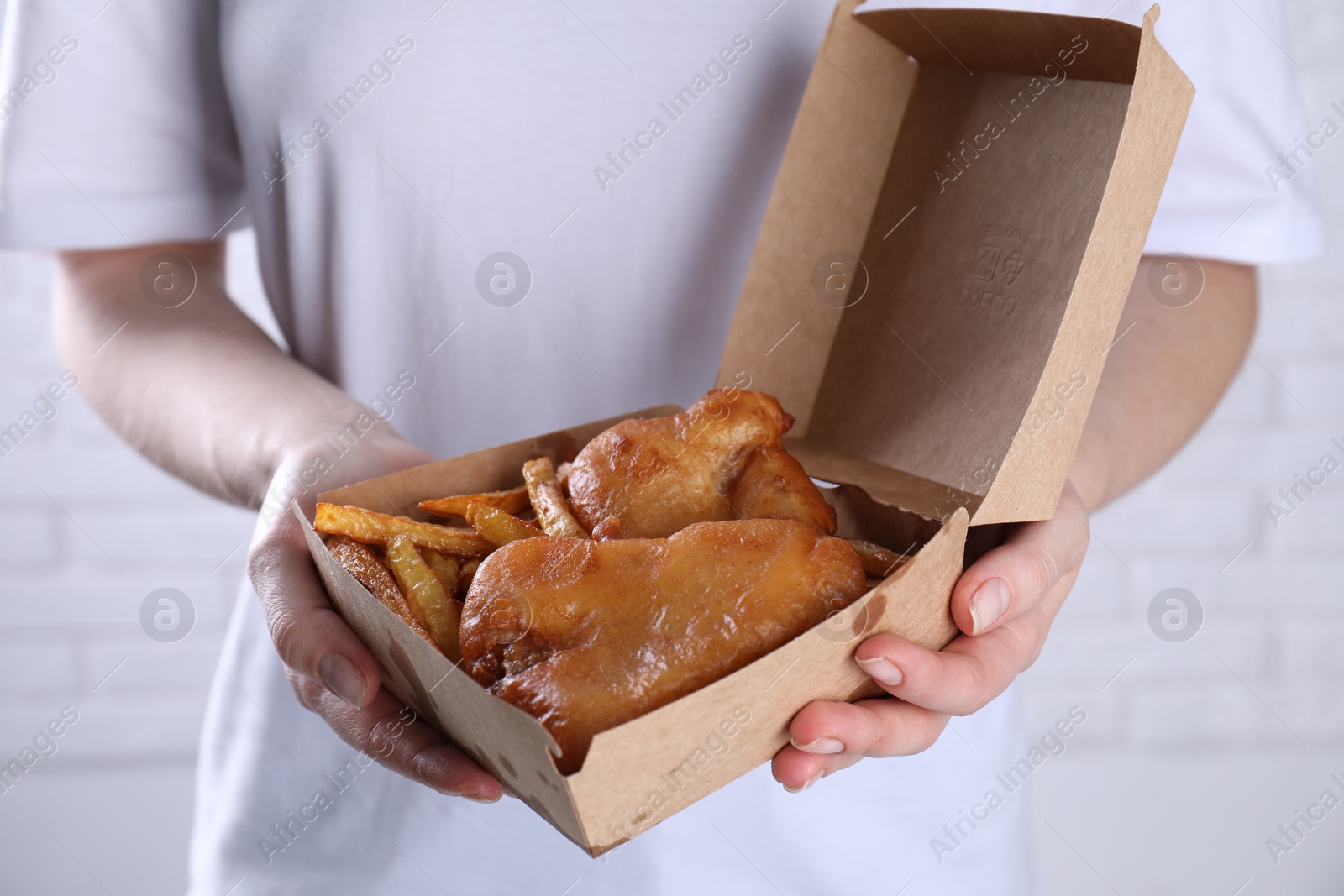 Photo of Woman holding fish and chips in paper box near white brick wall, closeup