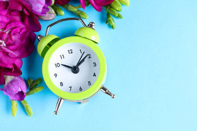 Green alarm clock with spring flowers and space for text on light blue background, flat lay. Time change