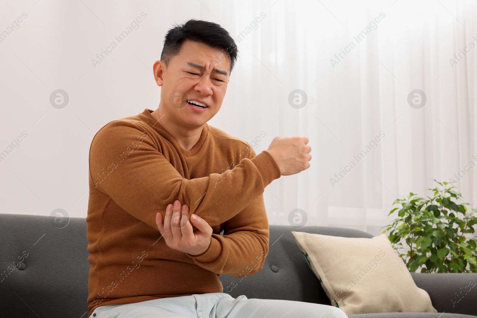 Photo of Asian man suffering from pain in his elbow on sofa indoors