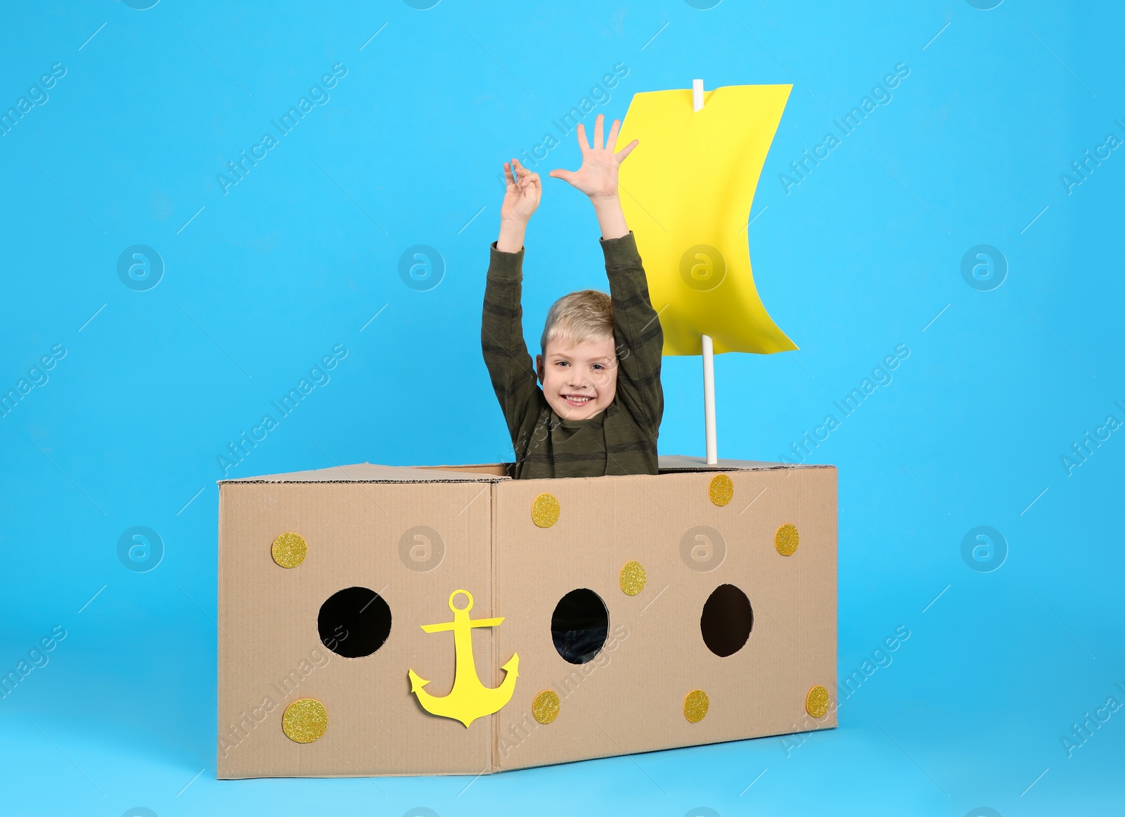 Photo of Little child playing with ship made of cardboard box on light blue background