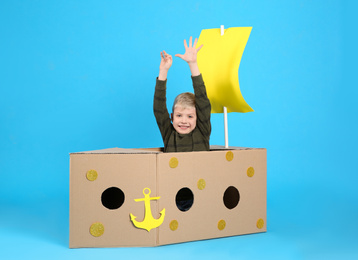 Little child playing with ship made of cardboard box on light blue background